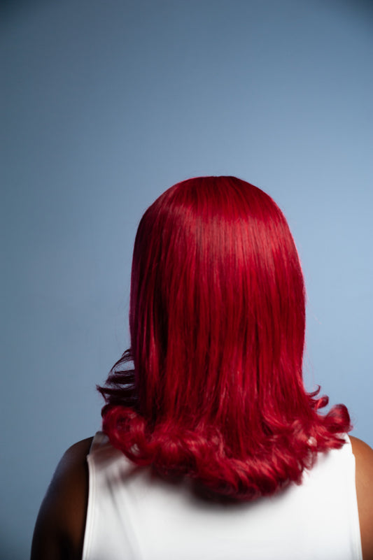A person shown from the back wearing a bright red shoulder-length wig with soft waves, against a solid blue-gray studio background.
